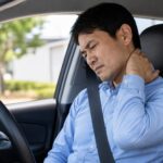 40代男性が車を運転中に寝違えによる首の痛みで困っている様子。振り向けず首を押さえているイメージ。